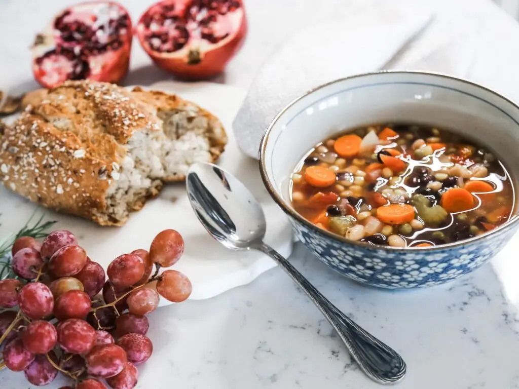 A white marble countertop holds an opened pomegranate, a cluster of purple grapes and a half-eaten loaf of crusty bread covered in seeds and oats. On the right is a small white bowl with a gold rim and white flowers on a blue background around the side. The bowl holds lentils, carrot slices, chopped celery and black beans in a clear broth. A metal spoon sits in the middle.