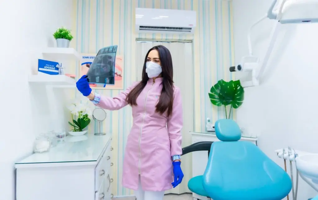 A woman with long brown hair and a pink zip-up medical jacket stands in a dental office. She wears bright blue gloves and holds up x-ray results. The office has white walls and one wall with stripes. The furniture is white and the seat is blue. Three indoor plants are in the background.