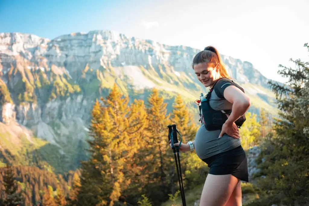 A woman in grey and black exercise closes stands on a high hill. She wears her long brown hair in a high ponytail and holds hiking sticks. A chest pack across her torso holds water bottles. She looks down smiling at her pregnant belly. A mountain is across the entire background behind her, plus the tops of some yellowing trees - how to prepare for labor