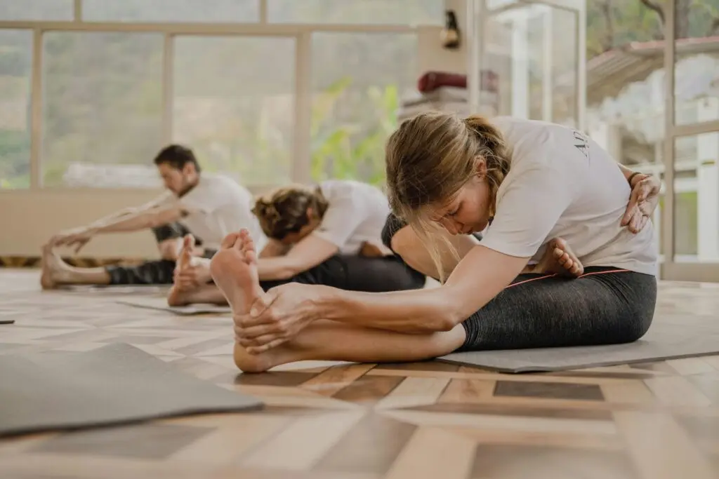 A group of people practicing yoga in a bright room with large windows and a nature view, performing a seated forward bend stretch on mats.