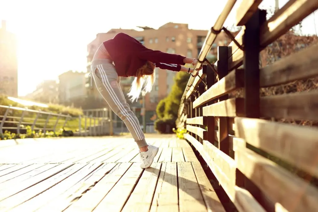A person in a red hoodie and gray sweatpants stretches against a railing on a sunny boardwalk.