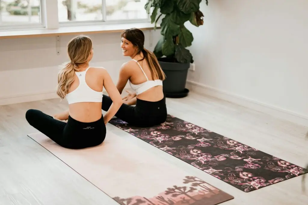 Two people in a yoga pose sitting on mats in a bright room with a large green potted plant. They are wearing white sports bras and black leggings, and are facing a window with sunlight streaming in.