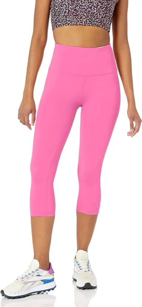 A woman stands in white tennis shoes with calf-length pink leggings that also have a high waist.