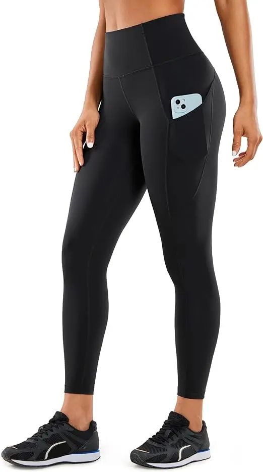 A woman stands in black tennis shoes and ankle-length, high-waisted black leggings. Her phone is in her pocket.