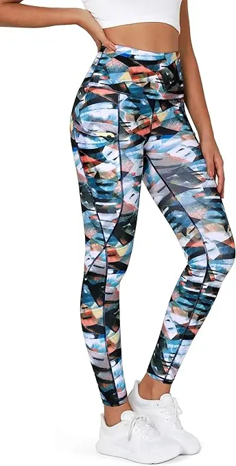 A woman stands with one hand on her hip while wearing white tennis shoes and multi-colored, high-waisted leggings that also have pockets.