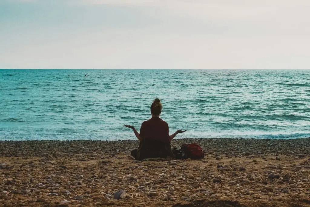 A woman sits cross legged on a rocky beach shore with waves stretching beyond her. She faces toward the water, away from the camera, with her palms up toward the sky and her hair in a bun. An orange bag sits on the rocky sand next to her.