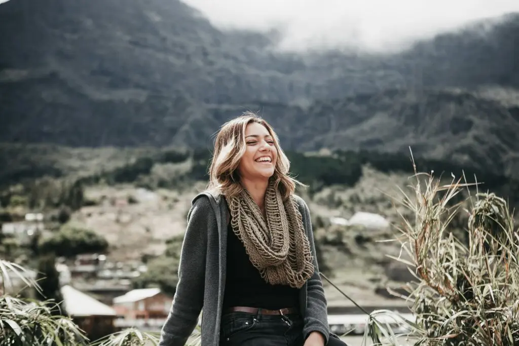 A woman wearing a black and grey outfit with a tan infinity scarf laughs with her eyes closed. Behind her is a small town on a hillside with cloudy mountains filling the image.