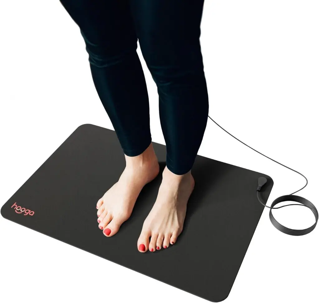 A woman stands on a black grounding mat with a black electrical cord against a white background and floor. Only her knees and feet are visible.