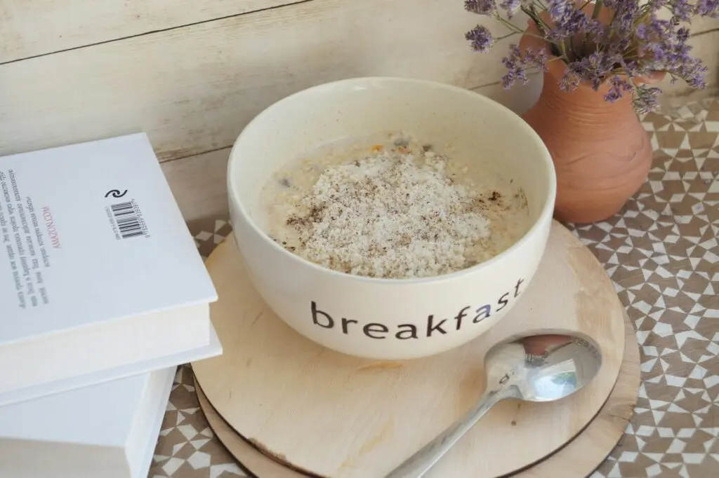 A white bowl that says "breakfast" filled with oatmeal. It sits on circular wooden place mats next to a silver spoon, two books and a clay pot of lavender flowers.