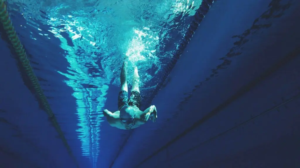 A man wearing a silver swim cap, reflective goggles and swim trunks pushes himself under water. There are lane markers floating above him.