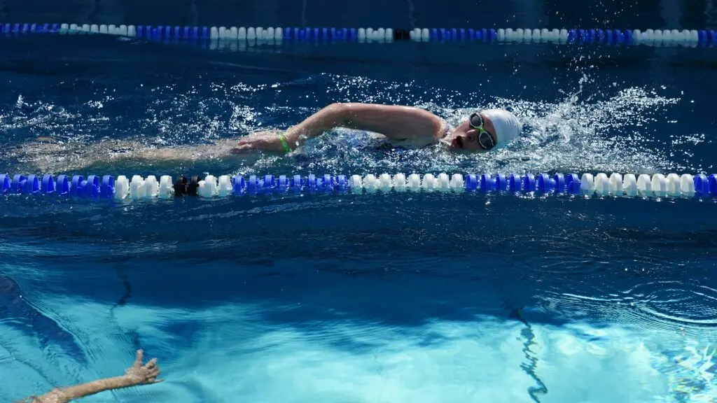 A swimmer in a white cap and clear goggles lined in black and green looks to the side to breathe while doing the freestyle stroke. The lane markers are a mix of blue and white. Someone swims in the next lane.
