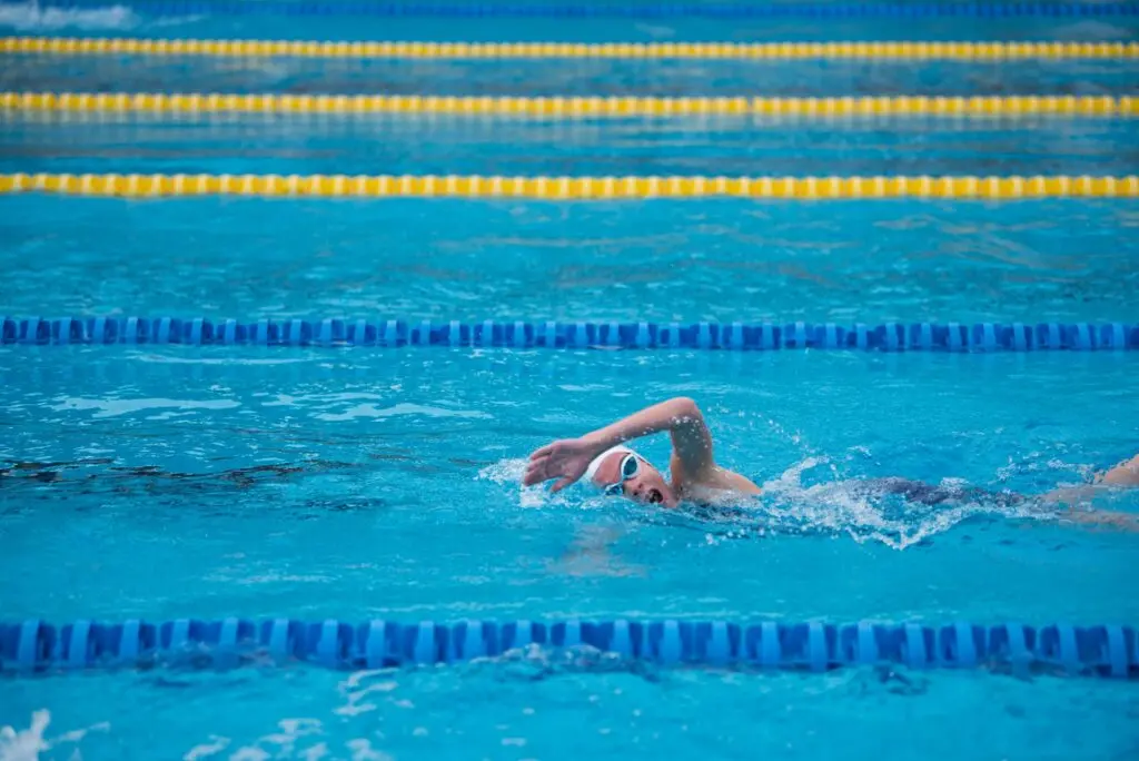A swimmer in a white cap and white reflective goggles lifts their arm during the freestyle stroke and turns their head to breathe. The lane markers alternate blue and yellow.