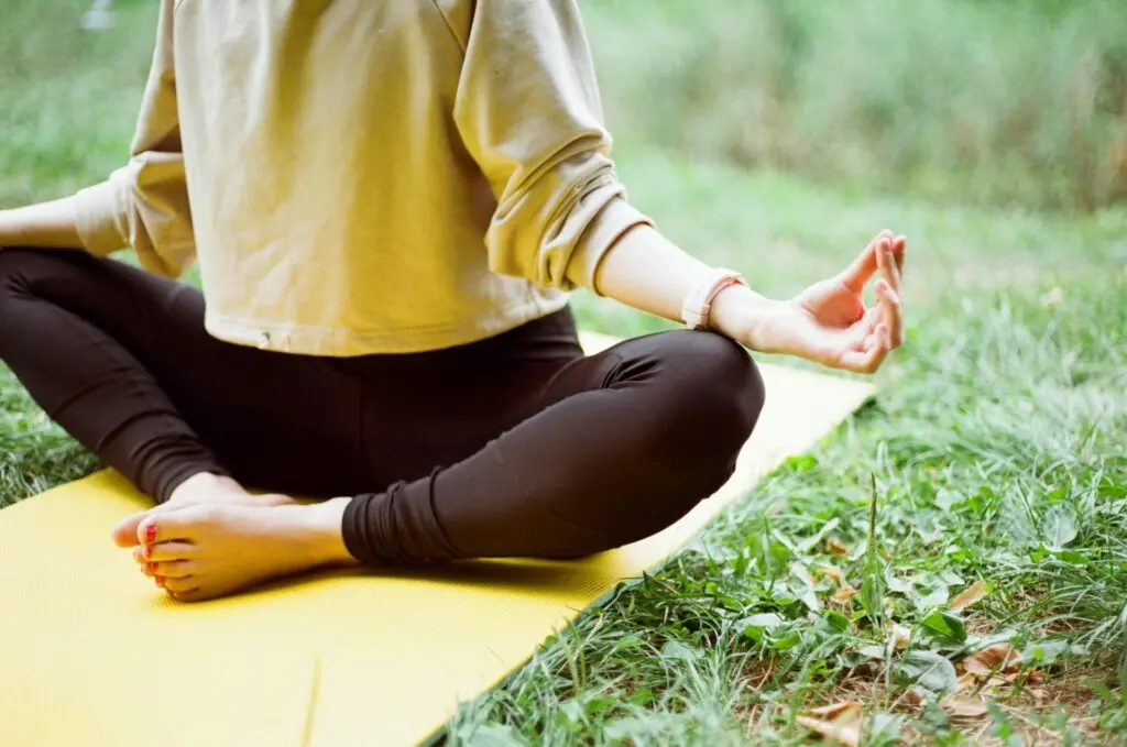 A woman sits on a yoga mat with her feet drawn in and touching. She holds her hands on either knee with her first three fingers touching. Green grass surrounds her yellow mat.