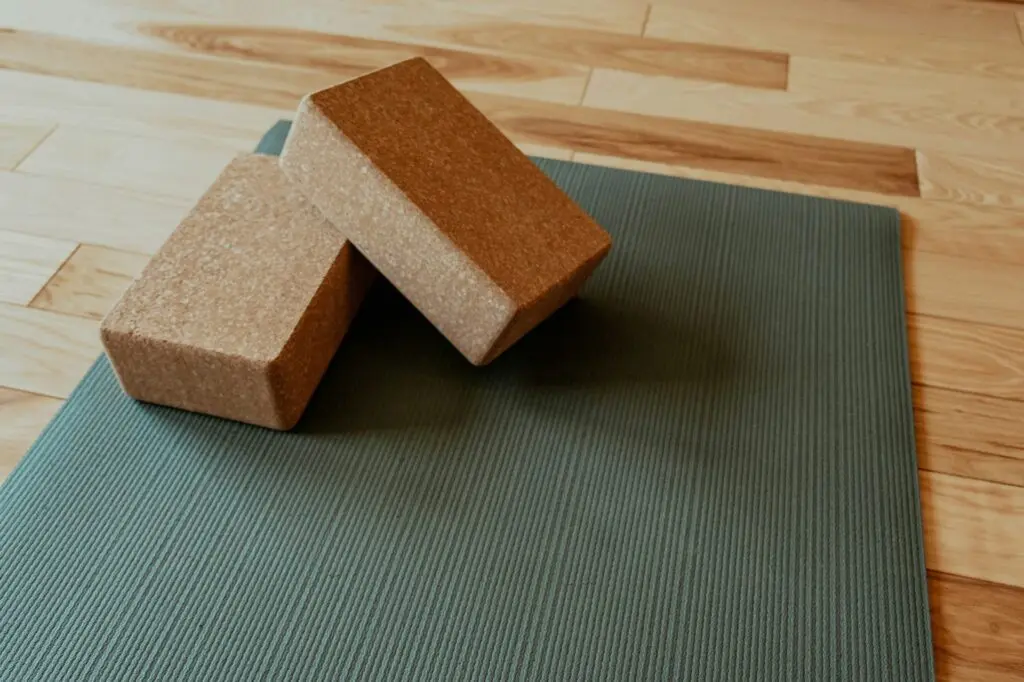 Two brown foam yoga blocks sit on a green yoga mat on light brown wooden flooring.