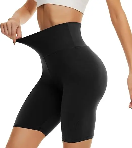 A woman in a white sports bra holds out the high waist of her elastic black workout shorts. They end at the mid-thigh point.