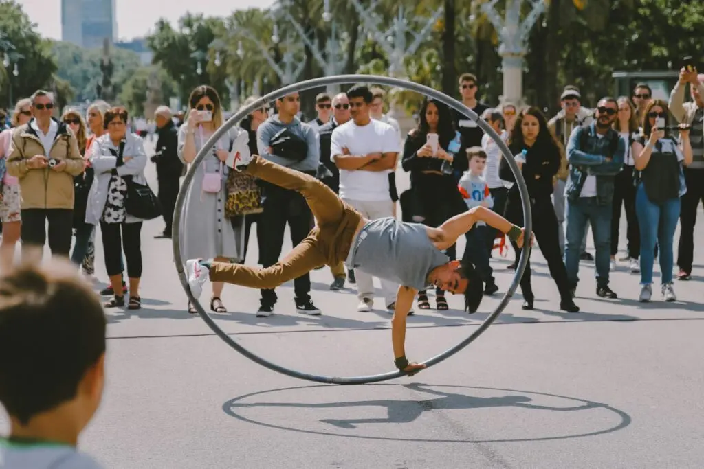 A man suspends himself in the middle of a human-sized hula hoop as part of a dance. A crowd watches around him because they're in a city park or street. Some people record him on their phones.