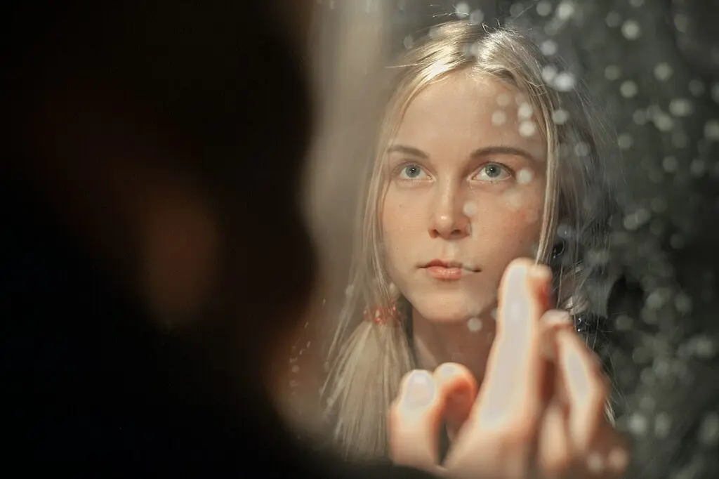 A blonde woman looks into a mirror covered in water droplets and touch it like she's trying to reach her reflection.