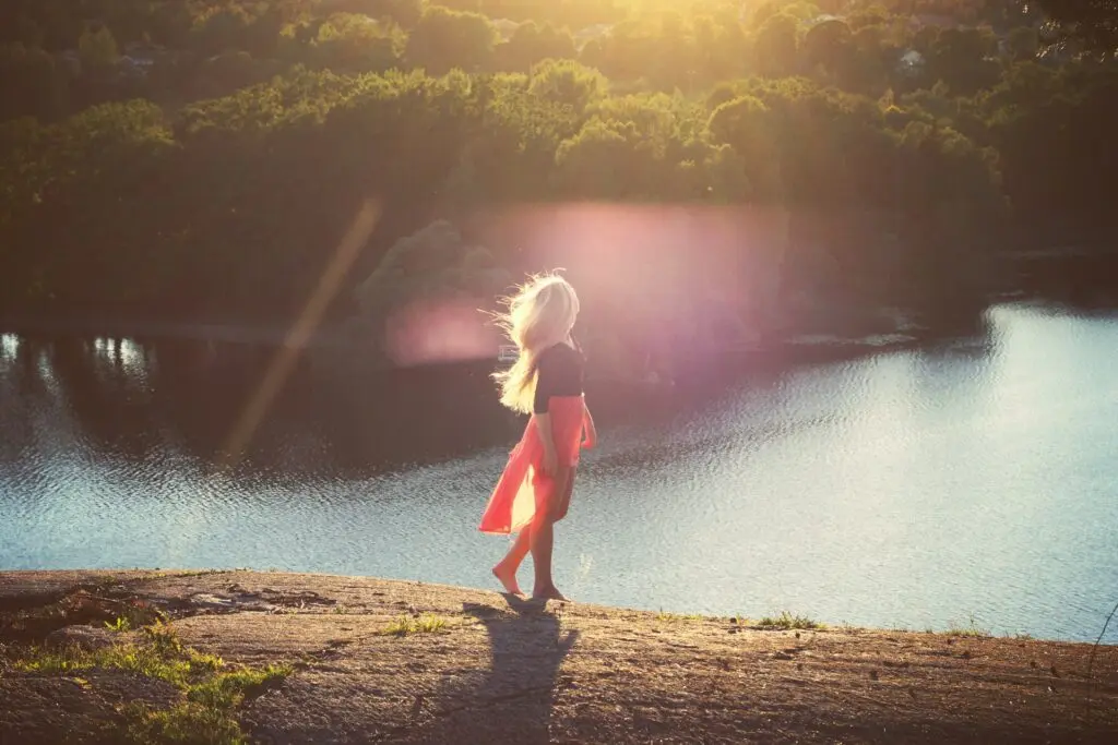 A woman uses one of the grounding techniques for dissociation by going for a barefoot nature walk under golden sunbeams by a river. Thick trees line the other side of the river.