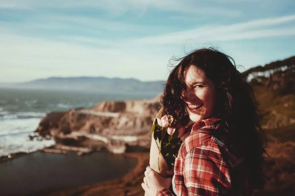 A woman stands on a cliffside by the beach, holding a bouquet of pink tulips wrapped in brown paper. She looks back at the camera smiling mid-laugh while the wind whips her hair in front of her face.