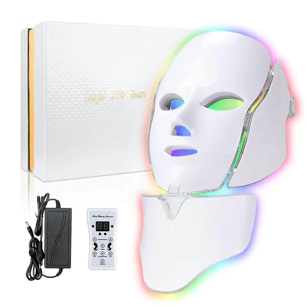 An LED light therapy mask sits against a white background with a box, a plug and a remote. It glows in multi-color around the edges of the mask.