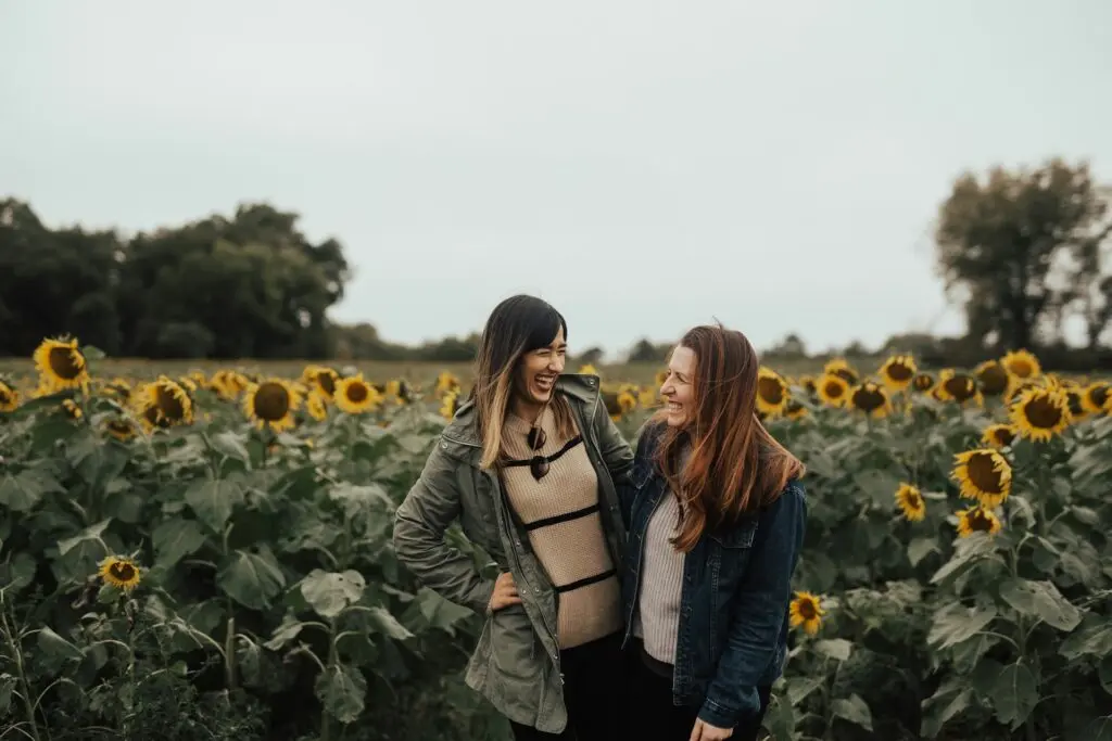 Two women laugh together in a sunflower field under a cloudy sky. They're both wearing light sweaters and jackets.