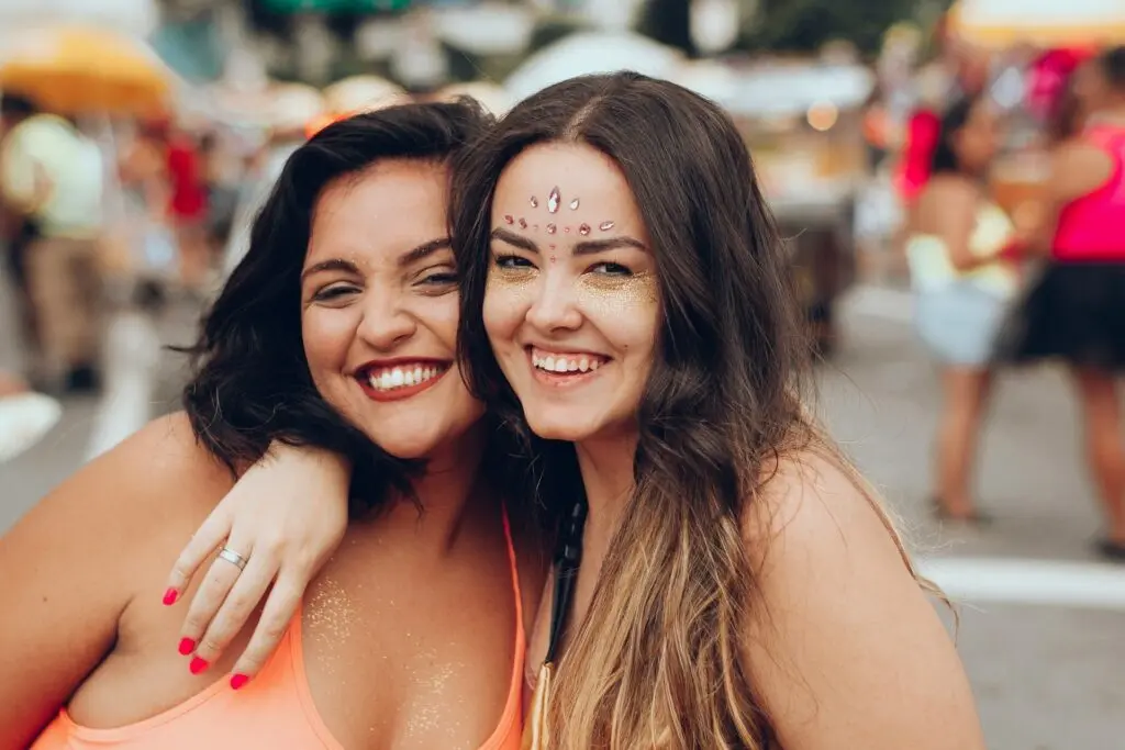 Two women hug each other and smile. They're at a summer festival and wear sleeveless shirts. One of the women has glitter over her chest and the other has it over her cheeks and her forehead is bedazzled. A crowd is blurred in the background.