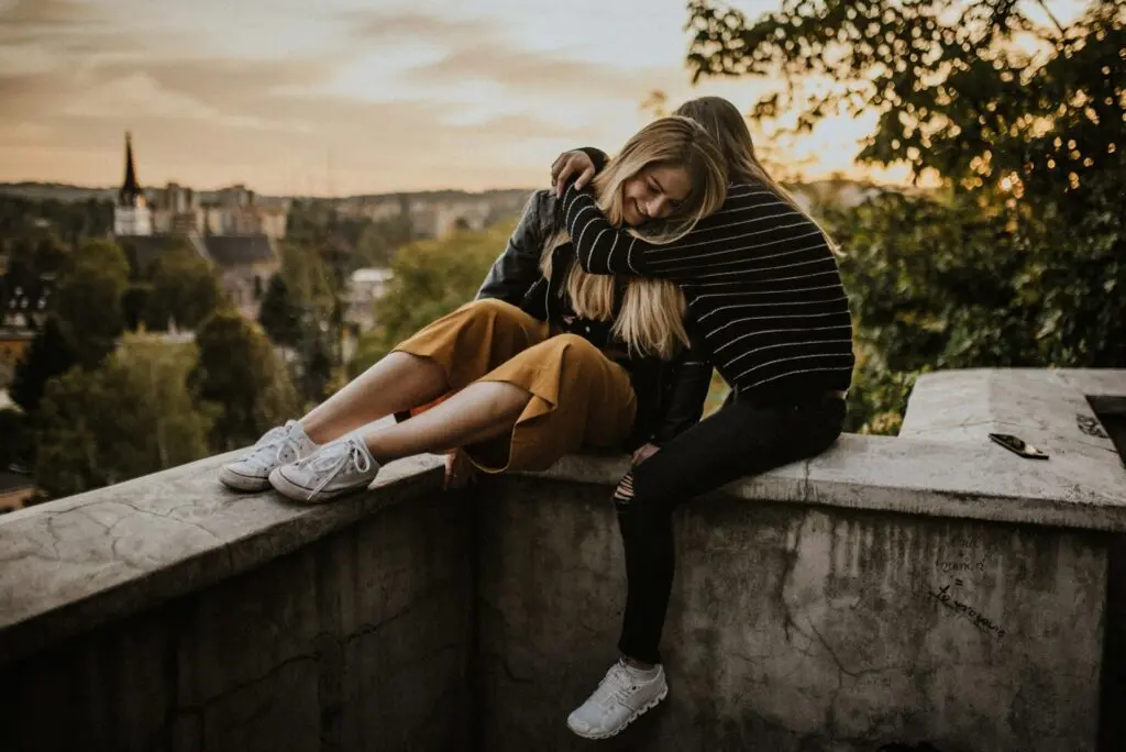 Two friends hug each other on a stone rooftop. An older, European city with a tall steeple stands in the background, surrounded by trees. The sun is setting in the background.