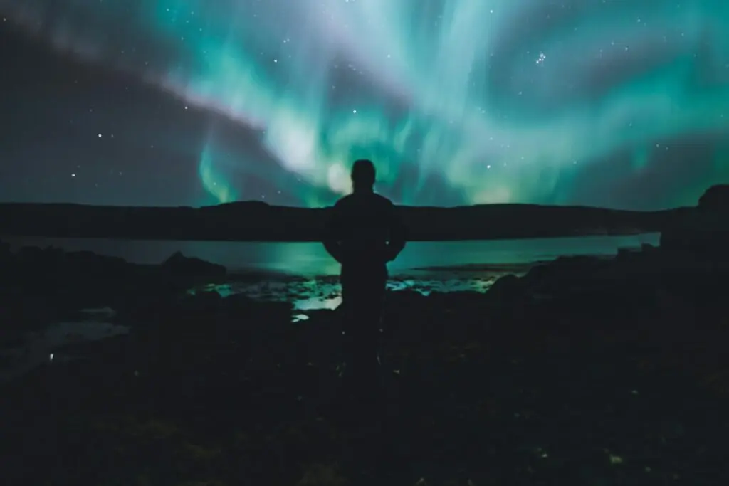 A night owl stands underneath the blue and green Aurora Borealis in the middle of the night. A lake is in front of him and he's on the rocky shore.