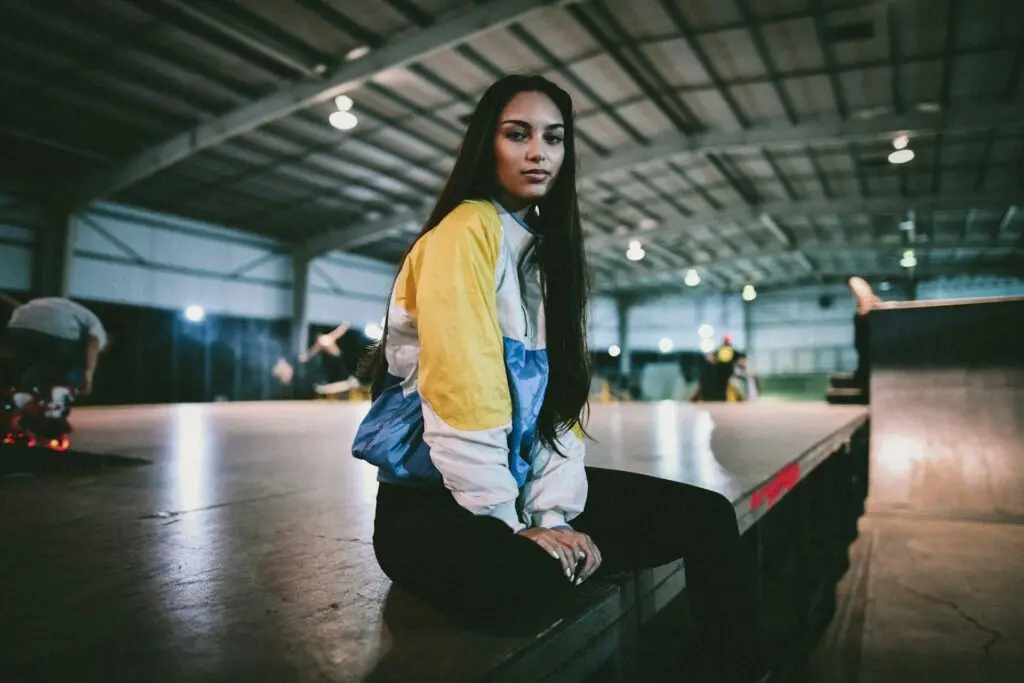 A woman with long dark hair sits on an empty stage in a warehouse. Two men set up equipment behind her. She's wearing a yellow, white and blue striped jacket and black leggings.
