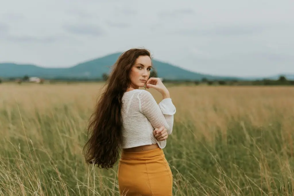 A woman with long brown hair looks back over her right shoulder. She's wearing a white long-sleeved crop top and a yellow pencil skirt. She stands in a wheat field at the base of distant green hills. The sky is full of light gray clouds.