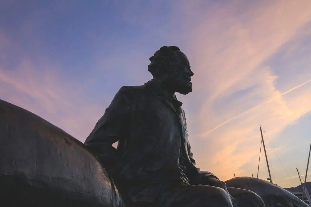 A picture of a statue of Jules Verne sitting on top of giant octopus tentacles. It's sunset and the sky is blue and pink above him.