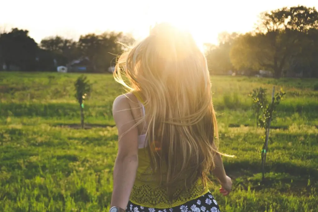 A woman with blonde hair flowing in the wind runs toward a green field with houses just beyond it. She wears a white and green tank top and the sun is setting in front of her.