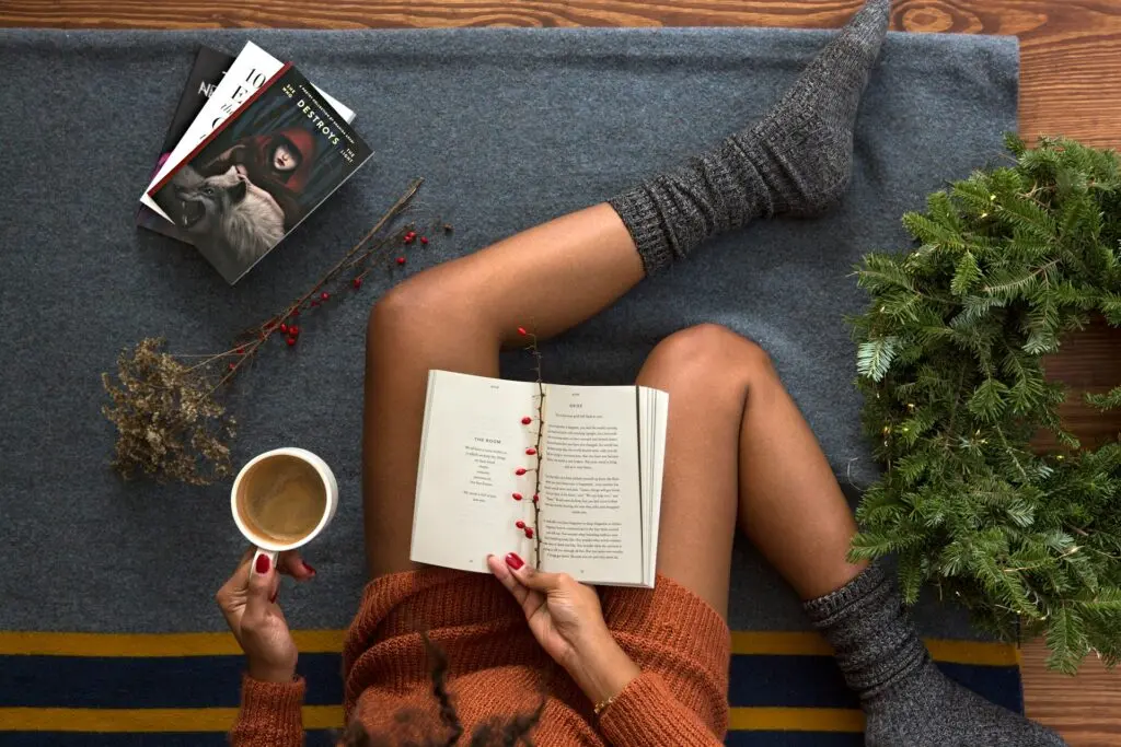 A woman sits in gray socks and an orange knit sweater dress on a gray, blue and yellow blanket on a wooden floor. She holds a novel open in one hand and a white mug of coffee in the other. Three books sit on the blanket in front of her, along with a dried branch of some kind of flower and a stem of red berries. An undecorated green wreath sits to her right. Her legs are hairless.