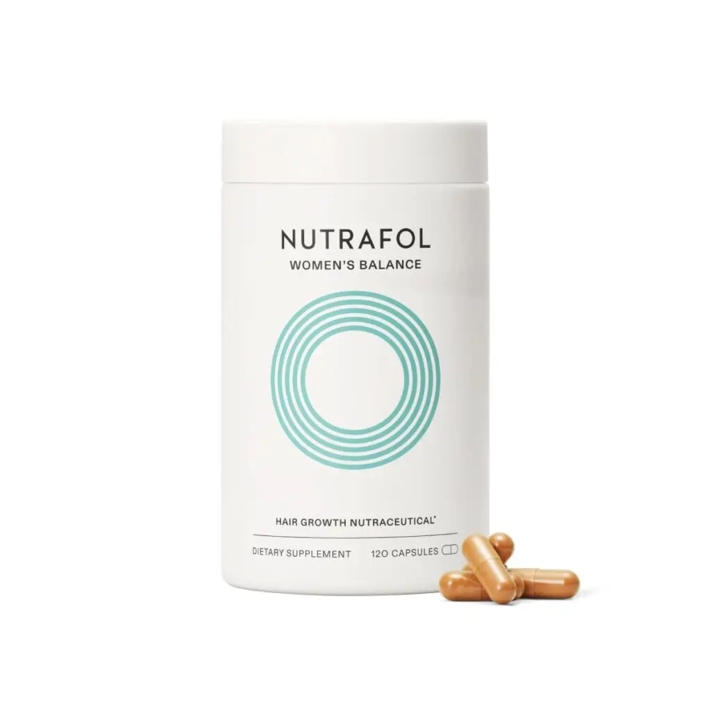 A white round bottle of Nutrafol hair growth nutraceuticals sits against a white background. Four brown capsules sit in front of it on the right.