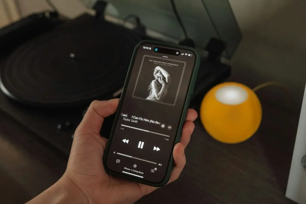 A person holds up their smartphone, which is playing "I Can Fix Him (No Really I Can)" from the The Tortured Poets Department anthology album. The background is an opened, empty record player and a round yellow speaker on a dark wooden dresser.