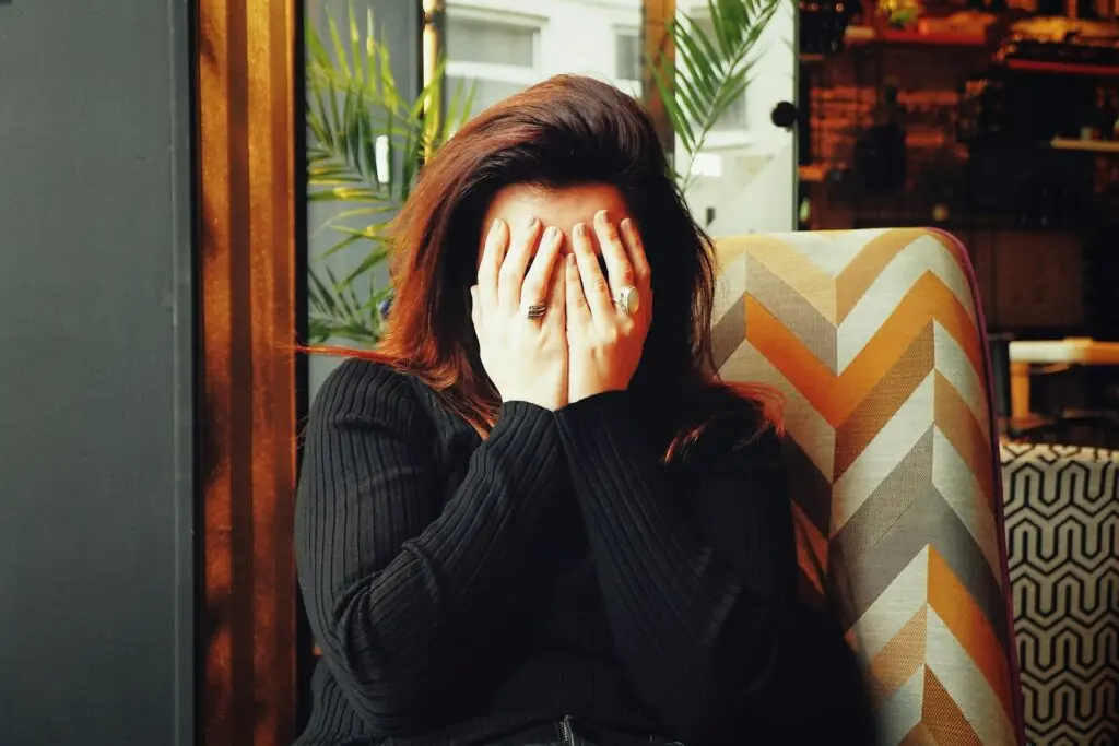 A woman with brown hair and a black sweater sits in a multi-colored chair in a room with lots of wood decor and plants. She covers her face with both hands because she's wondering if nausea after Pap smears is normal.