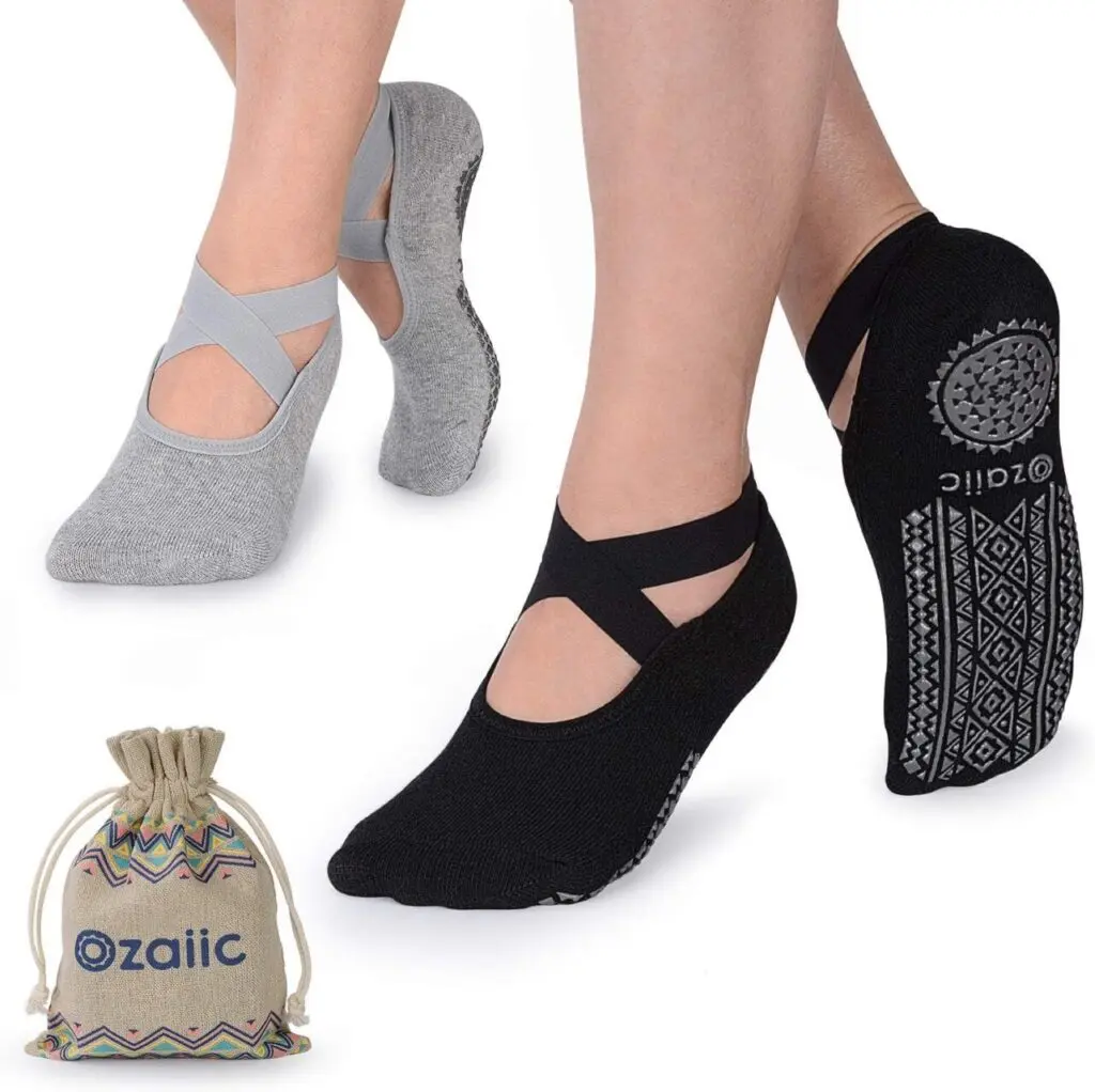 Two people each wear one pair of non-slip socks. The socks curve around the ankle and cross over the top of the foot. One pair is black and one is gray. The sticky patches on the bottom form geometric shapes and are gray. The knapsack they come in is in the bottom left corner.