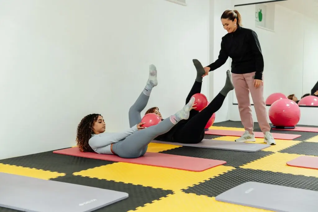 A woman dressed in pink sweat pants and a black long-sleeved shirt helps two women who lie on the floor. They each hold a pink ball between their legs as they bicycle kick in the air. The floor is made of foam puzzle pieces. A mirror lines the far wall. The other wall is white.