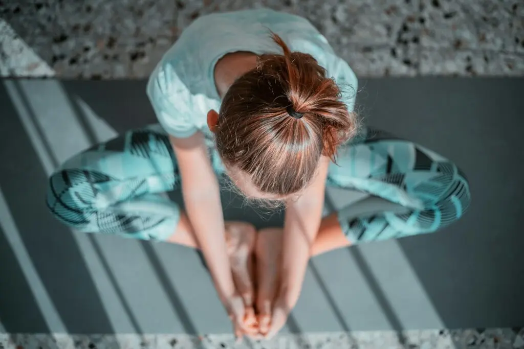 A woman in teal workout clothing sits with her feet pressed together on a teal yoga mat. She holds both feet in her hands and bends over them. The floor is speckled gray, black and brown beneath her.
