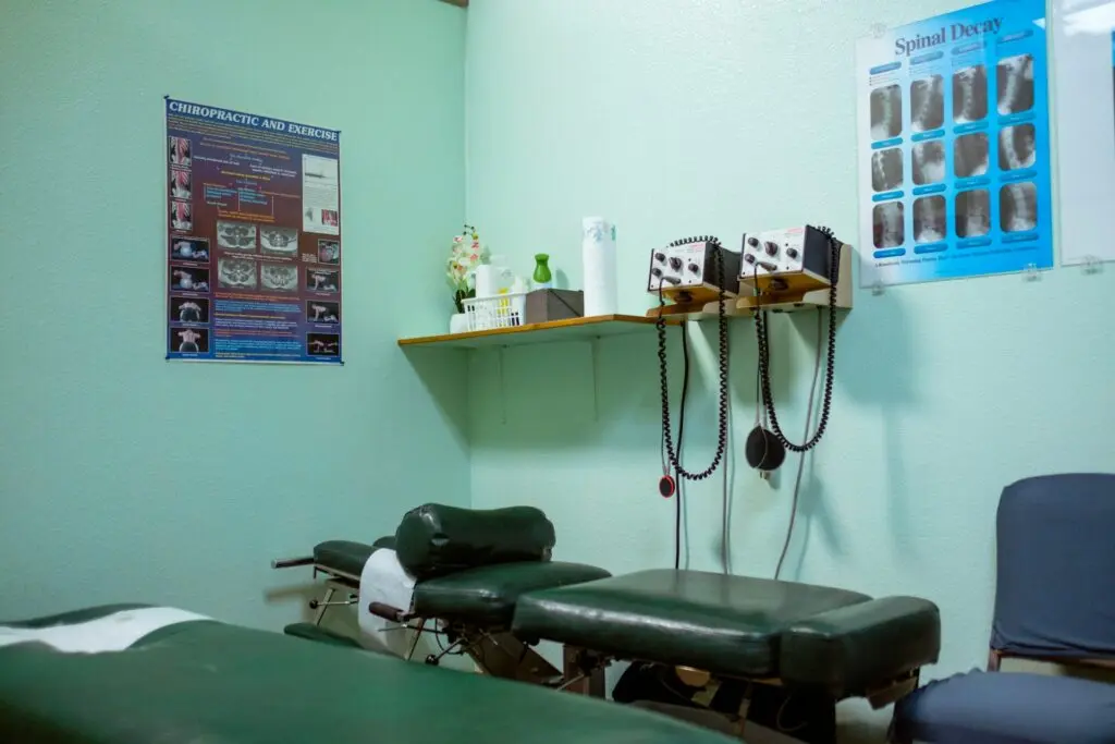 A chiropractor's office sits empty. The walls are blue-ish white and there are two green-ish tables next to a blue chair. A shelf contains some supplies and vase of faux flowers. Posters about chiropractic methods, exercise and the spine are on either wall.