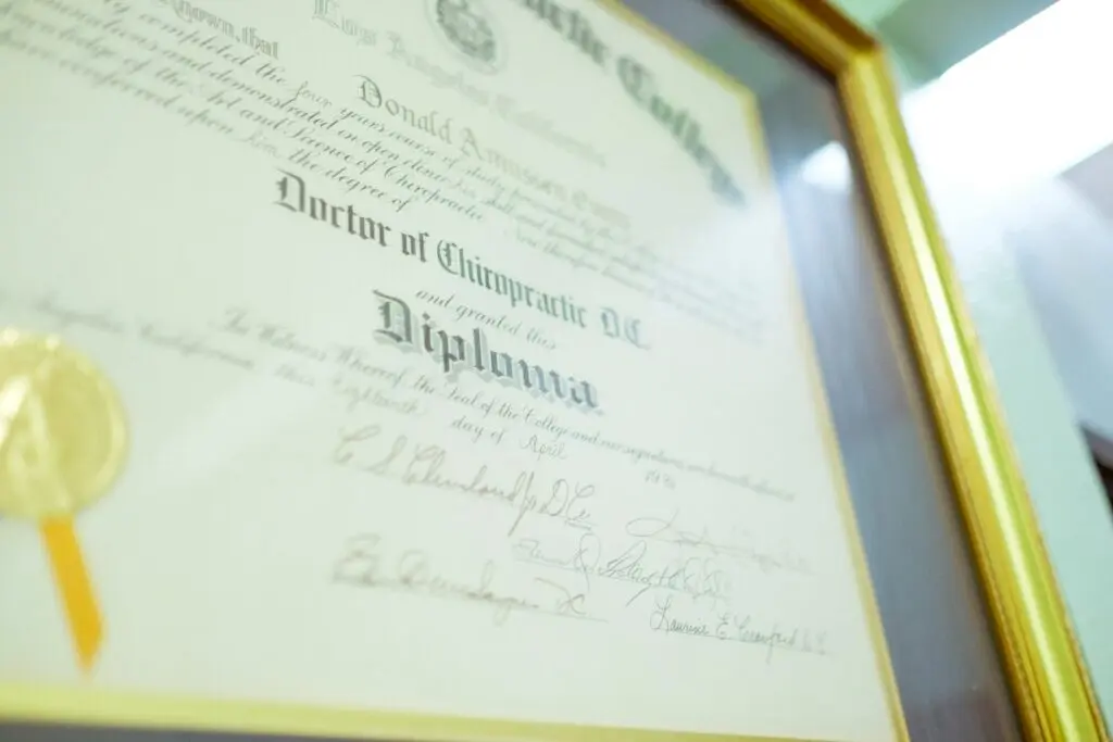 A man named Donald's doctor of chiropractic diploma sits in a gold frame on a wall within a room lit by florescent lighting.