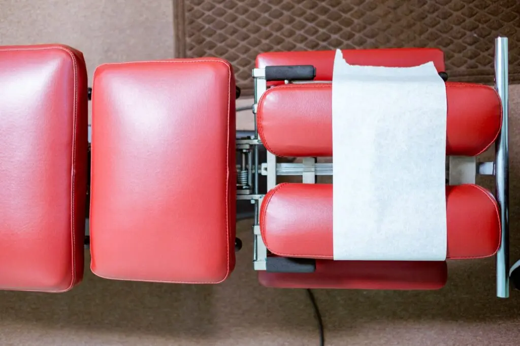 A red chiropractor's table with white medical paper over the headpiece sits in an office. The floor is brown marble and there's a brown rug in the top right corner.