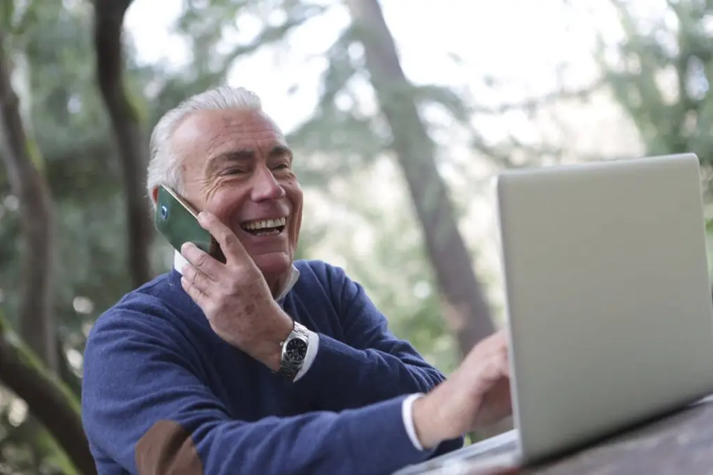 An older adult speaking on the phone in front of an open laptop