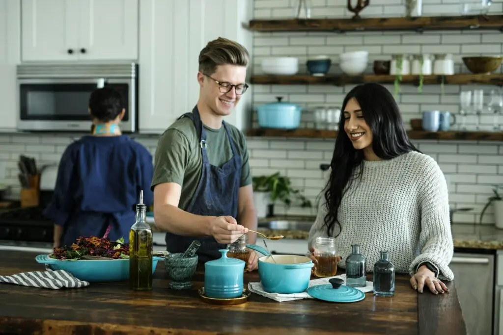 A man and a woman are making a recipe in light blue cookware on an island in their kitchen. The kitchen has modern interior decorating like white subway tiles, white cabinets and upcycled wood shelving. A third person cooks something on the stove behind them, like food that fits within their 7 day carb cycling plan.