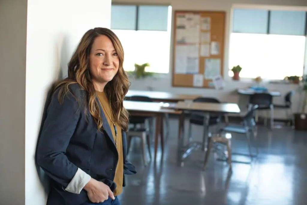 A woman in a gray blazer and tank shirt stands against a white wall in a grade school classroom. The desks behind her are empty.