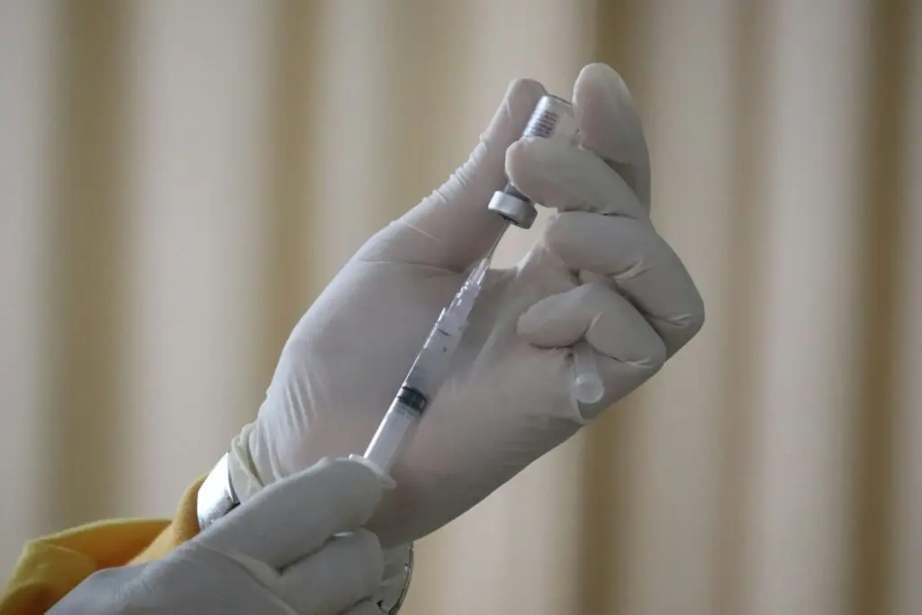 A person wearing white gloves inserts a needle and syringe into a glass vial. They hold the vial upside down while extracting the clear liquid with the syringe.