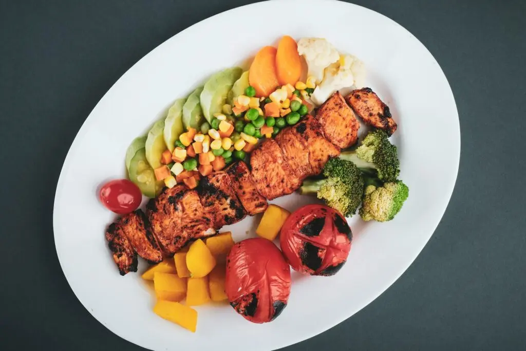 A white plate covered in vegetables, fruit and bite-sized, charred chicken pieces sit on a gray table for someone eating the paleo diet.