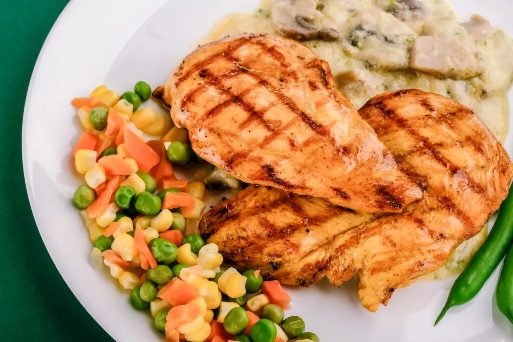 A white plate of chicken, mushrooms, corn, peas and carrots sits on a green table.