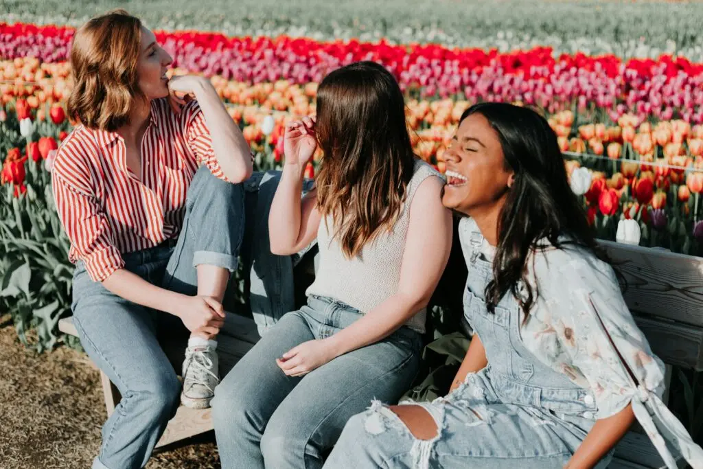 Three women sit and laugh on a bench in a field of red, orange, pink and white tulips.