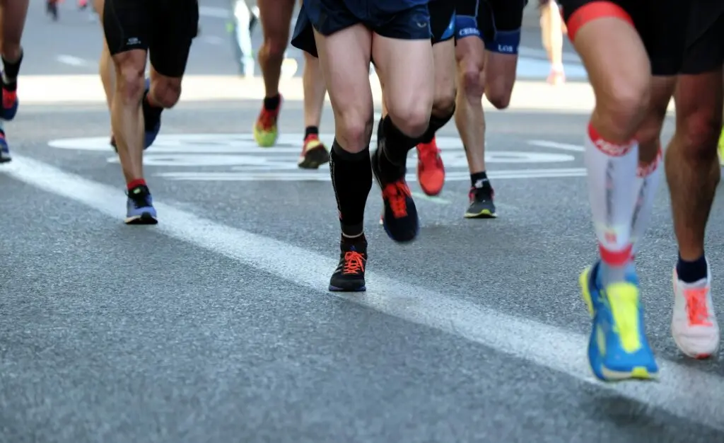Seven runners race across a road. Only their tennis shoes and legs are visible. Everyone is wearing multi-color clothes and shoes.